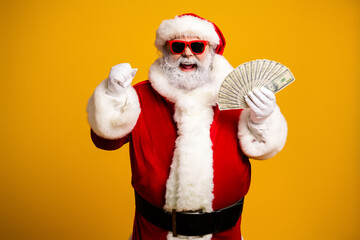 Santa Claus smiling with cash in hand wearing red costume and sunglasses against yellow background during holiday season