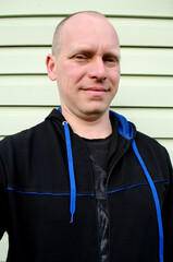 Casual smiling bald man wearing dark hoodie standing outdoors near vinyl siding
