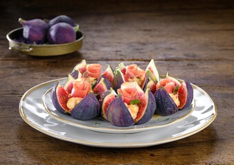 A plate presents figs, carefully sliced and filled with creamy cheese and cured meat. A small bowl...