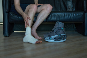 Person with injured foot sitting near orthopedic boot. Recovery phase. Bandaged foot and medical boot indoors.