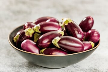A collection of small, shiny eggplants, with their green stems still attached, are piled high in a dark bowl. The bowl rests on a gray, textured surface