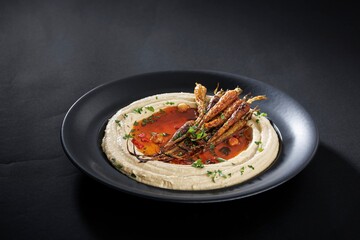 On a sleek, dark plate, hummus is swirled around, topped with roasted carrots, a vibrant sauce, and flecks of herbs, creating an inviting dish