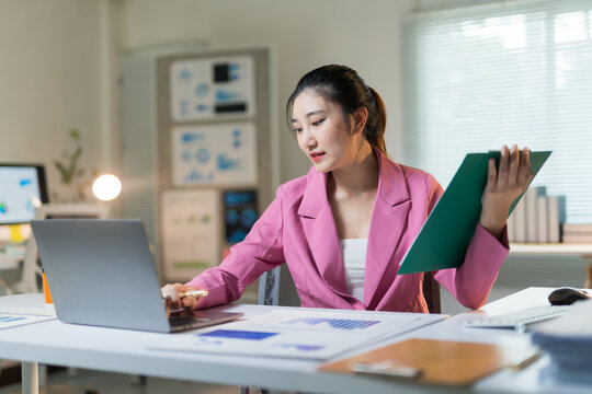 Asian business woman wearing a pink suit checking financial data on a laptop, holding documents, working in a modern office