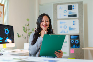 Asian businesswoman holding a clipboard, reviewing market data and financial charts with a happy, confident smile in her modern office
