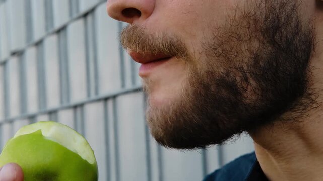 A man in a dark blue shirt eats a green apple while sitting near a modern metal wall on a sunny day. Close view. Horizontal 4k footage