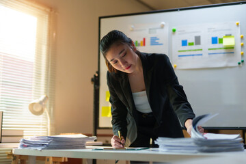 Asian businesswoman working at an office desk, writing on documents near stacks of paperwork, with...