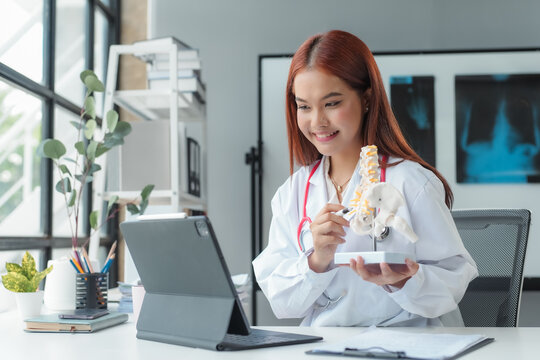 Female doctor providing telemedicine consultation, explaining human anatomy using a spine model on a tablet in an office