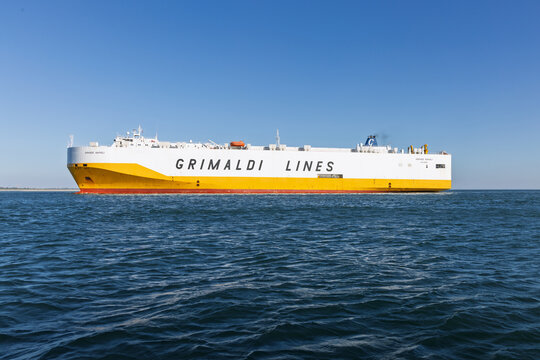 A large Grimaldi Lines roll-on/roll-off cargo ship (RoRo) sails on the blue water near Set&uacute;bal, Portugal. The distinctive yellow and white hull dominates the scene. Setubal, Portugal. 20 August 2025.