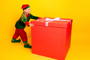 Cheerful young girl in festive elf costume with large red gift box against bright yellow background...