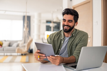 Smiling young man using tablet indoors wearing casual green shirt relaxing at home with laptop