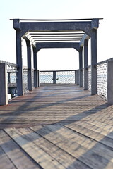 Fototapeta premium A wooden pier with a concrete pergola extends over calm blue water, offering a peaceful view of the sea and distant islands. The structure is framed with rope fencing and dried vines.