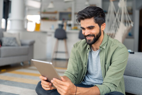 Smiling young man in green shirt using tablet in modern living room with laptop and coffee cup nearby