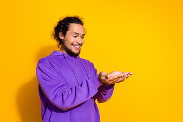 Portrait of a joyful young man in purple hoodie making hand gestures on vibrant yellow background