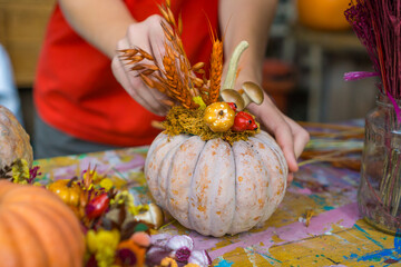 hands decorating a pale heirloom pumpkin with dried wheat stalks and ceramic mushrooms at a rustic workshop table, concept of diy craft, fall workshop, halloween, thanksgiving decor
