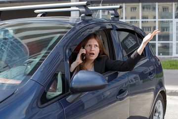 A beautiful young woman, with a phone in her hand, sits at the wheel of a car, gesturing and expressing emotions.