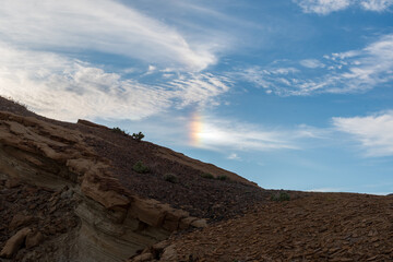 Layered rocky outcrop with shale and sparse desert shrubs under blue sky with wispy clouds and vertical rainbow, Death Valley National Park in April