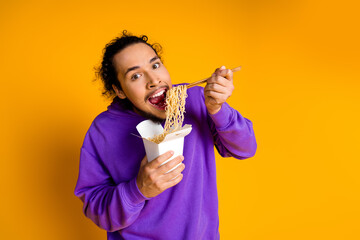 Portrait of a joyful man eating noodles from a takeout box against a vibrant yellow background...