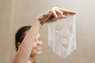 Happy woman holding a sheet face mask in a bright bathroom, enjoying self-care and beauty routine. Wellness and skincare concept.