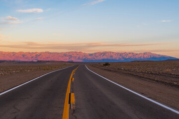Naklejka premium Desert Road Leading to Pink-Lit Mountains Under Pale Blue Sky with Pink-Tinged Clouds, Death Valley National Park