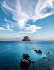 Coastal landscape with a solitary island.  Vast expanse of calm, azure water.  Dramatic clouds fill the sky.  Dark-toned rock formations near the shore