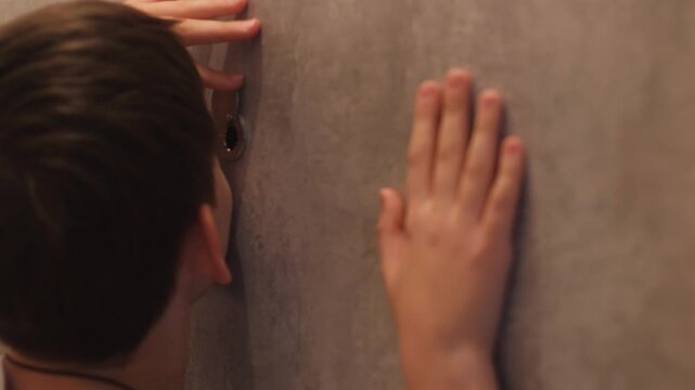 A Caucasian boy aged 10-12 years, being alone at home, looks through the peephole with apprehension, the child looks through the peephole