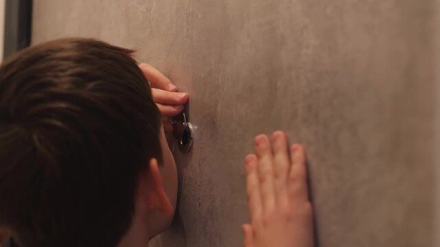 An 11-year-old Caucasian boy looks through the door's peephole while inside the house. The child, concerned about safety, looks through the door's peephole.