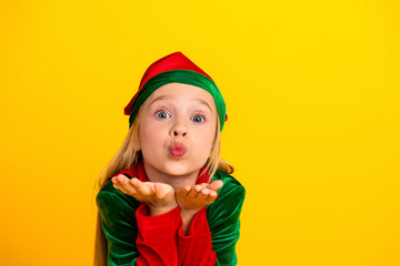 Cheerful young girl in festive elf costume blowing kisses against a vibrant yellow background celebrating Christmas magic