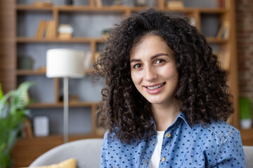 Smiling young woman with natural curly hair and brown eyes poses confidently in a cozy modern living room, radiating authenticity, warmth and relaxed professional charm