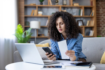 Woman reviewing household finances on laptop and phone, calculating bills and receipts in living...