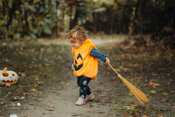 A young child in a pumpkin costume sweeps with a broom on a forest path during Halloween, with a carved pumpkin nearby