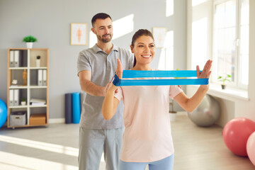 Happy smiling young woman patient doing physical rehabilitation exercises with elastic band under control of professional physiotherapist doctor. Health care, fitness therapy, physiotherapy concept
