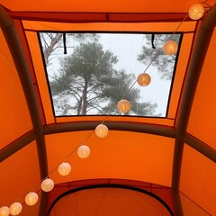 Fototapeta premium Looking up through an orange tent opening at a cloudy sky with trees and glowing string lights
