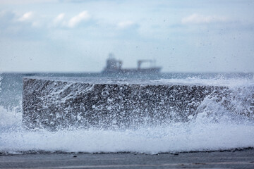 Waves crash on the stlny wall, in the background sea and ship in soft focus