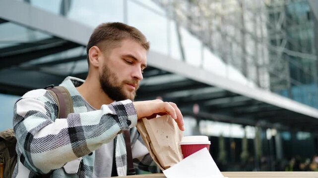 Young man with a beard and backpack enjoys eating traditional Serbian burek outdoors near a modern glass building