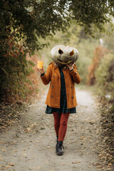 A woman in an orange coat and plaid dress with a carved pumpkin mask poses in an autumn forest on Halloween.