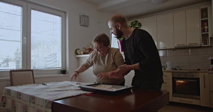 A senior lady prepares a meal with her son. They spread the dough on a baking sheet and put it in the oven.