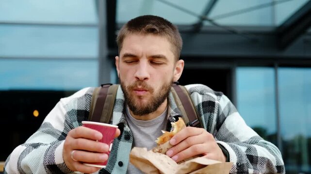 Young man with a beard and backpack enjoys eating traditional Serbian burek and drinking coffee outdoors near a modern building