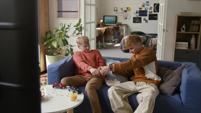 Wide angle shot of two teenage boys as brothers fighting for TV remote while sitting on couch at home watching TV together, copy space