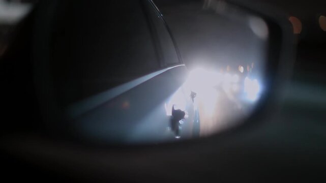 Bright headlights from vehicle behind seen in rear view mirror during night drive, extreme glare, abstract light shapes, traffic danger, disorienting urban scene