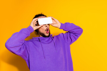 Young man in a purple hoodie interacting with smartphone against a yellow background showing excitement