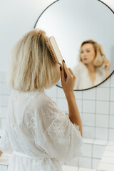 Back view of beautiful young woman in white undershirt combing her hair and smiling while looking into the mirror in bathroom