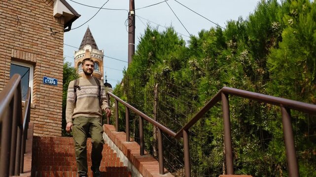 A man with a backpack go down on brick stairs on a narrow street in Zemun, Belgrade, with the historic Gardos Tower in the background
