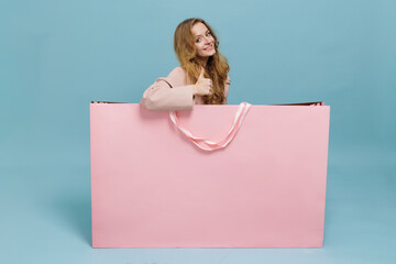 Woman giving thumbs up behind giant pink shopping bag on blue studio background