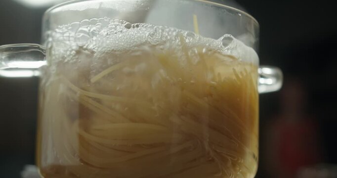 Pasta is cooking in a glass pot, steam rising above. Cook stirs with a wooden spatula hot water boils in a glass saucepan with spaghetti inside, steam gently rising. Transparent cookware cooking pasta