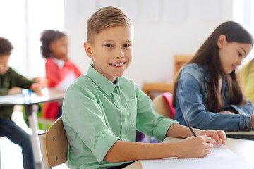 Obraz premium Positive European schoolboy sitting at desk in classroom during lesson in school and smiling at camera, diverse classmates on background