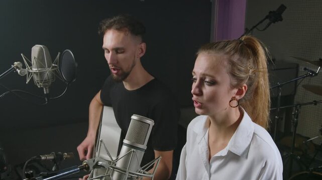 A man and woman collaborate on a musical recording, with the man playing guitar and singing while the woman sings into a microphone, creating a harmonious duet in a professional studio setting