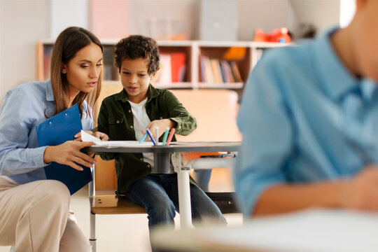 Young female teacher assisting Latin schoolboy, helping with task in test. Education, learning, childhood, teaching and school concept - Powered by Adobe