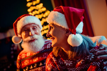 Joyful elderly couple in festive sweaters wearing Santa hats share a warm moment at home beside a...