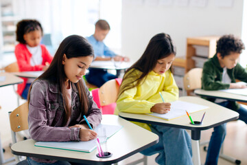 Smart diverse children sitting at desks and writing in copybooks, creating studious atmosphere in classroom interior in modern private school