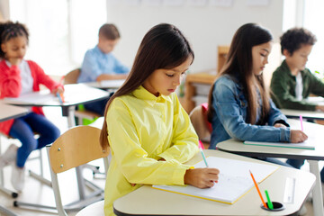 Focus on smart concentrated junior schoolgirl writing in copybook, having exam, children creating studious atmosphere in classroom interior